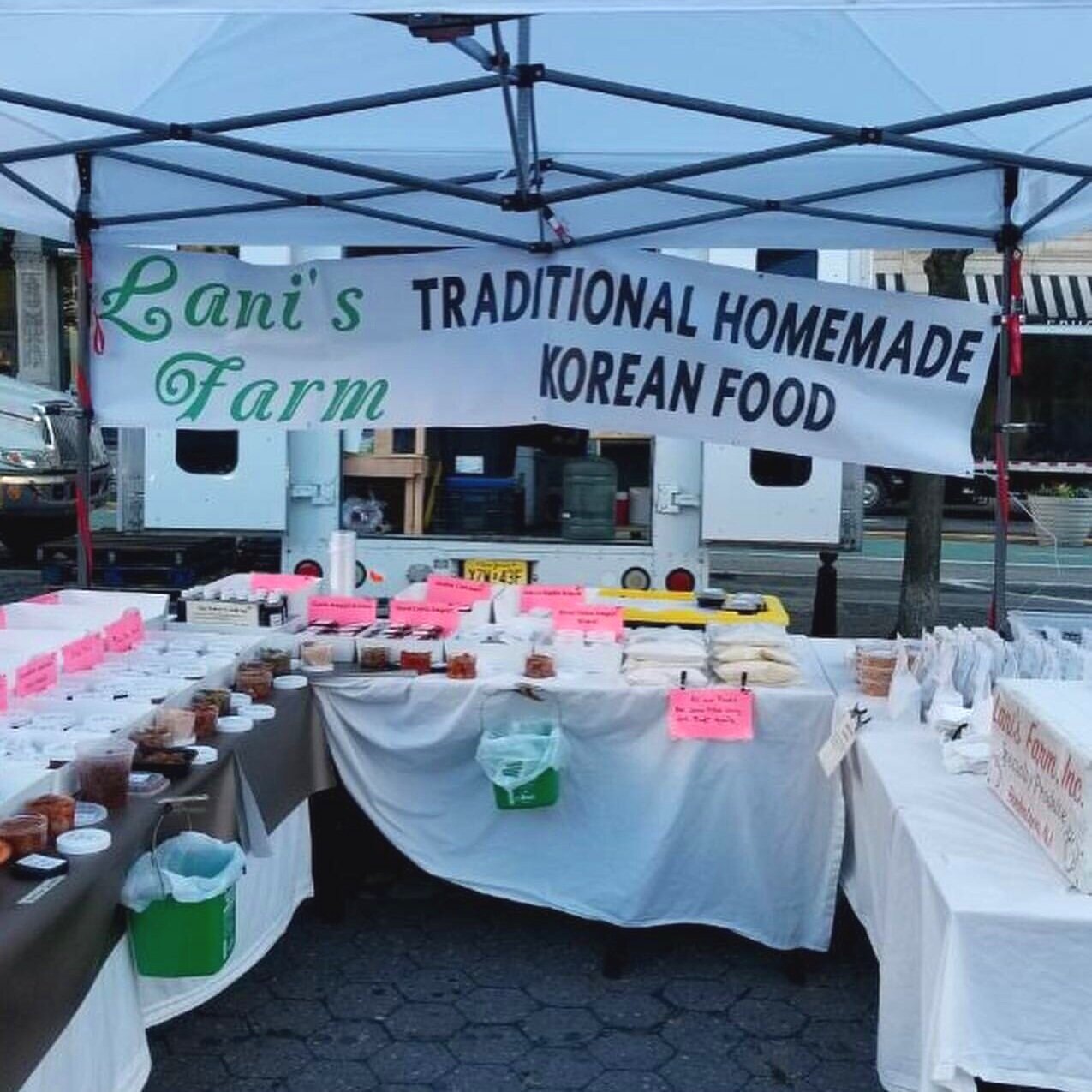 Lani's Farm booth at a market showcasing a variety of traditional homemade Korean food items, including jars and containers on tables with bright pink labels, under a canopy.