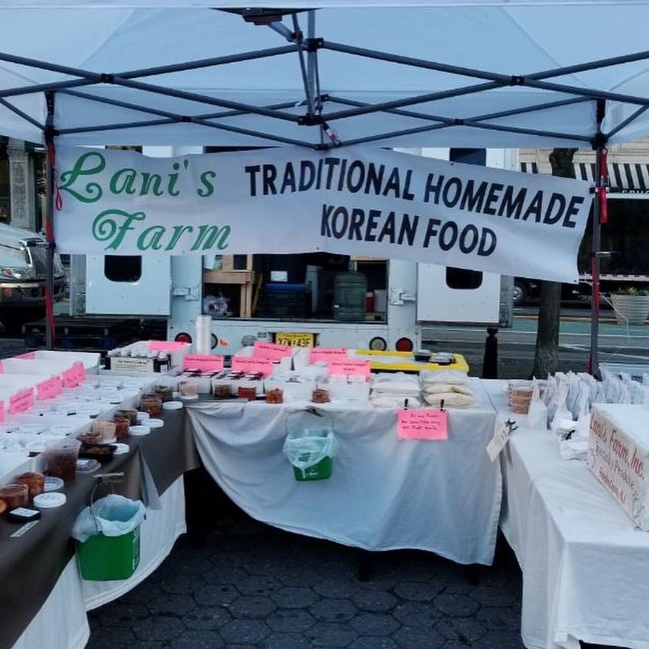 A vibrant market stall showcasing Lani's Farm, featuring a banner that reads "Traditional Homemade Korean Food." The table is filled with various jars and containers of Korean delicacies, labeled with bright pink tags, under a white canopy.